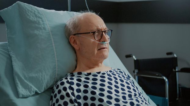 Portrait of patient with heavy breathing problems sitting in bed at hospital ward. Sick old man with nasal oxygen tube expecting doctor to receive medical treatment, medicine for illness