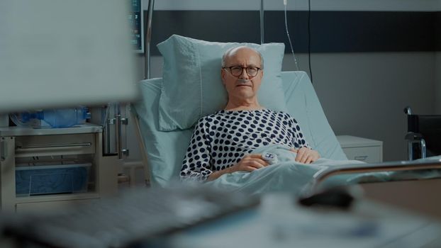 Portrait of elder patient sitting in hospital ward bed with nasal oxygen tube and oximeter to combat illness and health problems. Sick old man waiting for effects of healing treatment