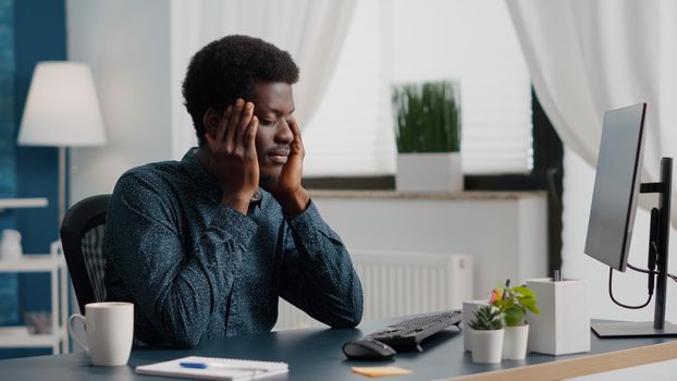 Stressing african american man with big headache pain, suffering from migraine, rubbing his temples. Overwhelmed manager working from home office, exhausted guy from problems and job related stress