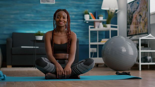 Portrait of black athlete sitting in lotus position on floor enjoying morning workout in living room. Woman in sportwear stretching muscle during strech gymnastics warming body