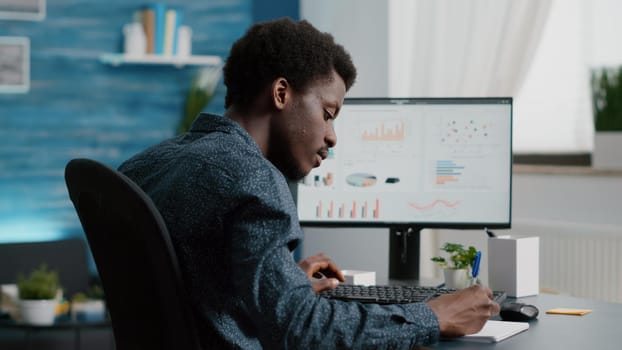 Selective focus on black american guy working from home, taking notes on notepad while remote working on computer from living room. Computer user using business internet online web communication job