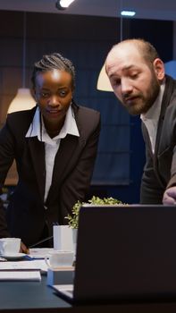 Diverse teamwork checking management presentation on laptop computer brainstorming company ideas. Multi ethnic businesspeople lean on conference table in office meeting room late at night