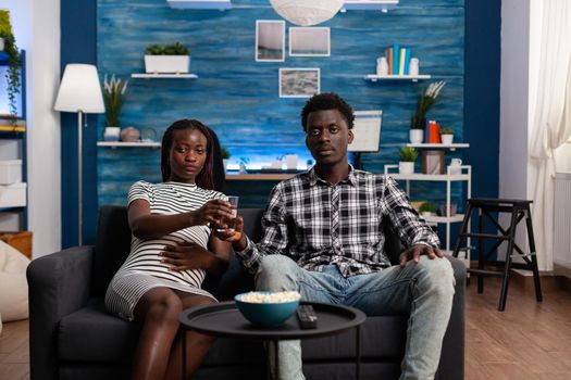 POV of black couple expecting child watching TV in living room. African american young family looking at camera and television while pregnant woman holding glass of water and hand on belly