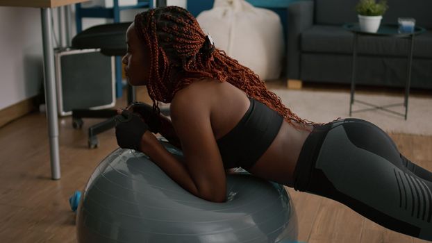 Athletic slim black woman stretching abdominal muscle while sitting on yoga fitness swiss ball during wellness morning workout in living room practicing sport. Fit adult doing aerobics position