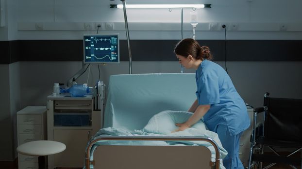 Medical nurse organizing blanket and pillow on hospital bed in clinical ward for treatment. Emergency room designed for recovery patients, surgery, illness, disease healthcare help