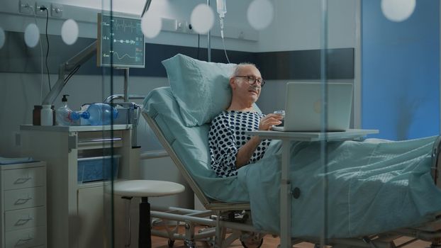 Patient in hospital ward talking on videocall with family using laptop. Sick old man with breathing problems wears nasal oxygen tube and oximeter while operating on modern computer