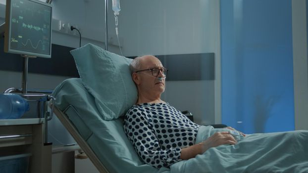 Sick patient sitting in hospital ward with nasal oxygen tube for heavy breathing problems. Old man with illness waiting on healthcare system to receive medical treatment and recovery