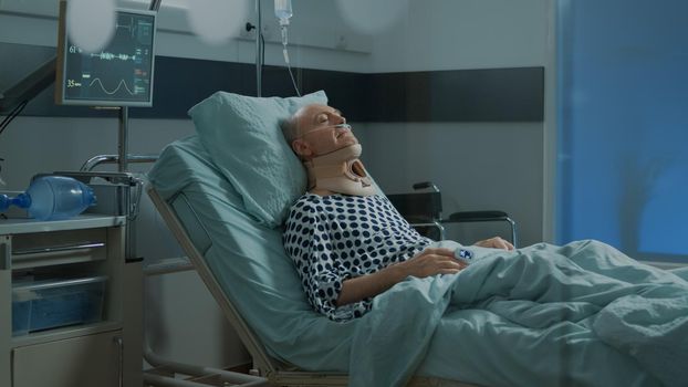 Old patient with cervical neck collar laying in hospital ward bed at medical facility. Sick elder man with health problems waiting on recovery from injury in modern intensive care unit