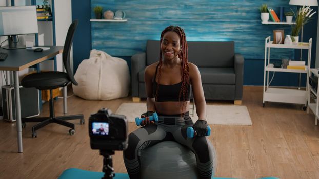 Black woman trainer practicing morning yoga exercise sitting on fitness swiss ball in living room recording aerobic training using dumbbells. Athletic working body muscles enjoying healthy lifestyle
