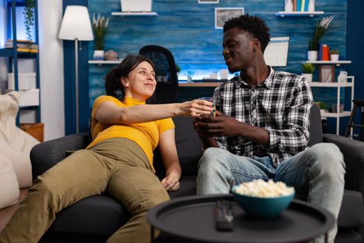 Interracial people with pregnancy sitting at home. African american father of child bringing glass of water for pregnant caucasian woman laying on couch. Mixed race couple expecting baby