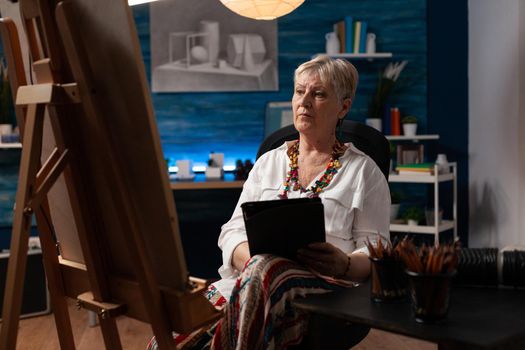 Elderly woman holding tablet and seeking inspiration for art masterpiece in creative workspace. Old artist using device and technology for drawing creation on canvas at professional studio