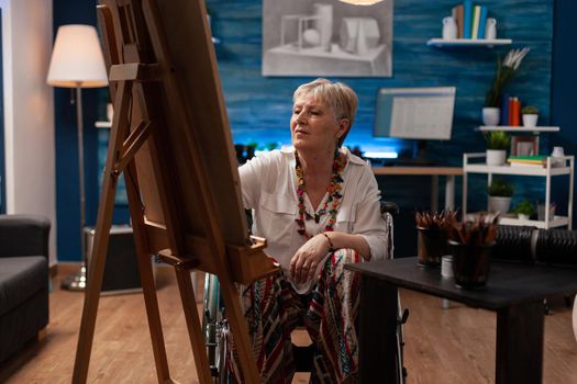 Elder artist with disability drawing on white canvas with easel at studio. Senior woman sitting in wheelchair while using pencil for professional masterpiece in workshop space