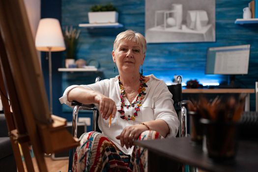 Portrait of disabled old artist sitting in artwork studio at home. Elder woman with handicap in wheelchair preparing for masterpiece drawing as hobby. Senior person with disability