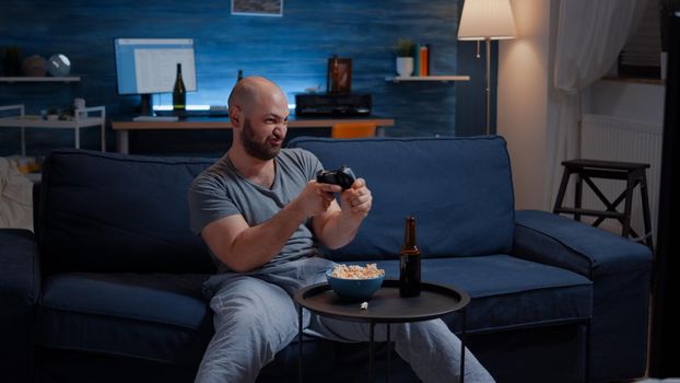 Determinated excited man sitting on sofa playing video games, happiness scream enjoying online competition. Competitive gamer using controller joysticks playstation gaming and having fun winning