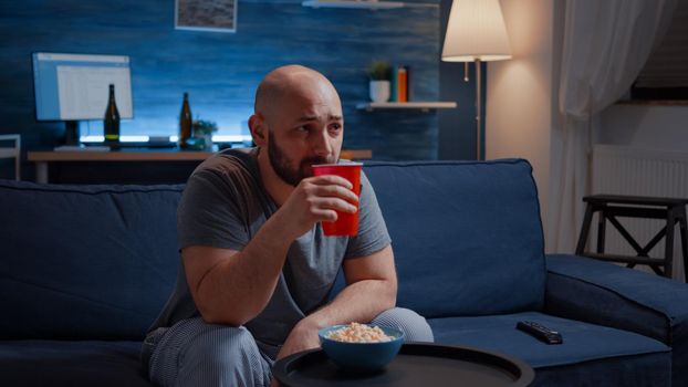 Man enjoying time watching tv series at home sitting on comfortable couch dressed in pajamas eating popcorn. Excited amused home alone male drinking juice on cozy sofa in living room late night