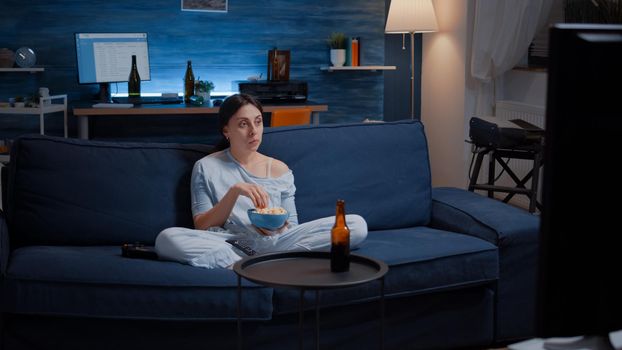 Young woman eating popcorn and watching an interesting serial on tv. Focused concentrated female home alone at night with surprised face looking at suspense movie sitting on comfortable couch