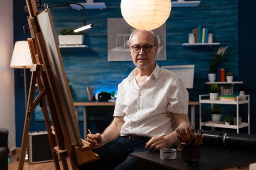 Portrait of caucasian elder artist sitting in artwork studio. Senior man with artistic imagination looking at camera while working on vase drawing. Aged person with masterpiece on canvas