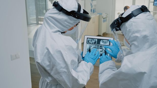 Orthodontists at dental clinic holding digital tablet with x ray scan of dentition for modern oral consultation. Stomatologists wearing ppe suits examining teeth radiography on device