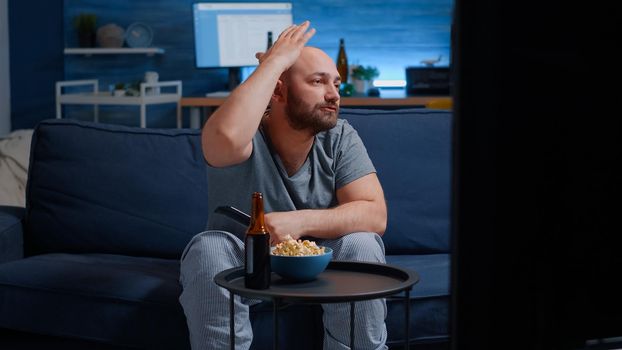 Upset man football supporter watching sport game supporting favourite team eating nervos popcorn after losing championship. Dissapointed male gesturing talking alone and drinking beer