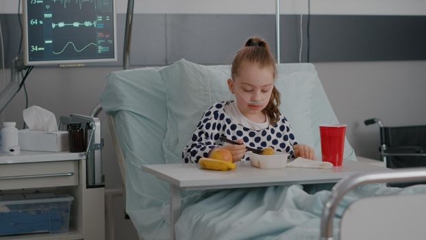 Portrait of hospitalized little child resting in bed eating healthy food meal during recovery examination after suffering medical surgery. Sick kid wearing oxygen nasal tube having breakfast nutrition