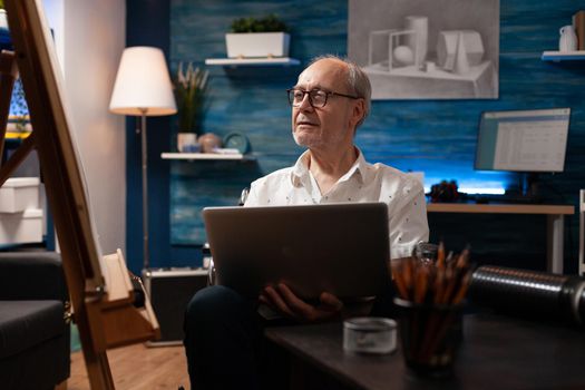 Caucasian man with handicap using laptop for drawing sketch in artwork studio. Elder artist sitting in wheelchair while working on professional masterpiece and holding modern gadget