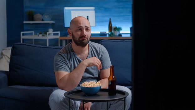 Sad guy watching drama movie on TV crying wiping eyes sitting on couch alone at night looking at television, eating popcorn. Sensitive person affected by film action, emotionally awestruck reacting