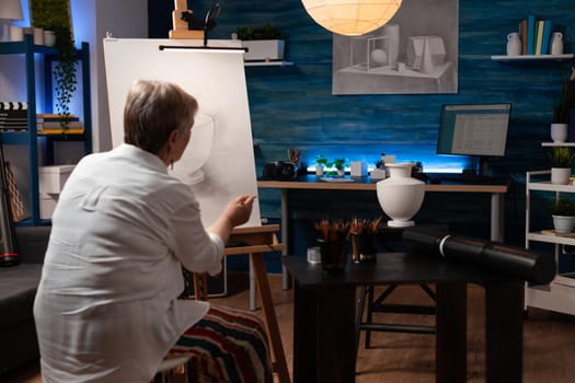 Senior artist sitting in artwork studio drawing vase design on canvas and easel using pencil. Old caucasian woman working on authentic masterpiece with object on table at workplace