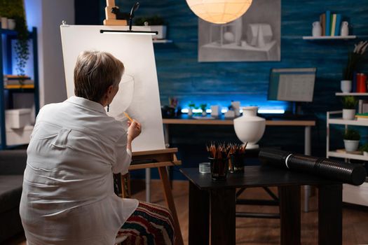 Caucasian aged artist working with vase and pencil on canvas for authentic drawing at workshop. Senior woman with artistic hobby creating masterpiece for fine art project at studio