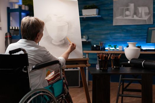 aged artist drawing vase on canvas using pencil in artwork studio. Senior woman with handicap sitting in wheelchair while creating professional masterpiece for fine art