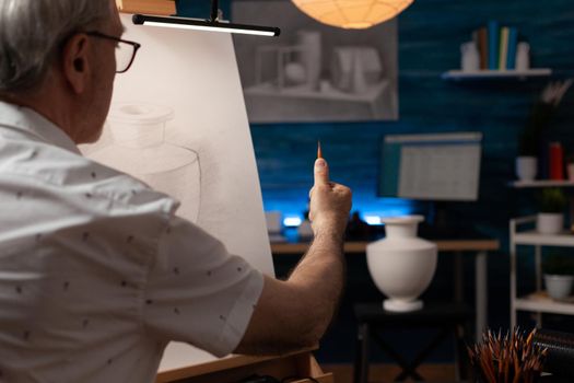 Caucasian senior man drawing vase design on white canvas in fine art studio. Elderly artist creating professional masterpiece on wooden easel in artwork space. Old adult with artistic hobby