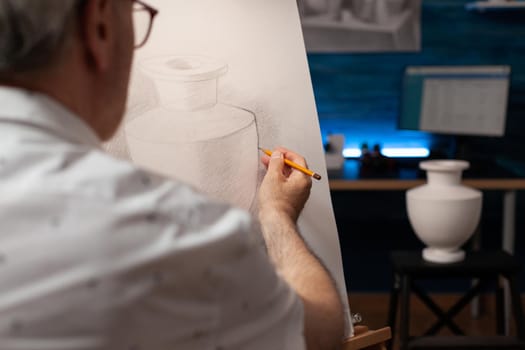 Caucasian artist looking at vase on desk and drawing on canvas at workplace. Senior man with artistic hobby recreating professional masterpiece using art intruments and tools.