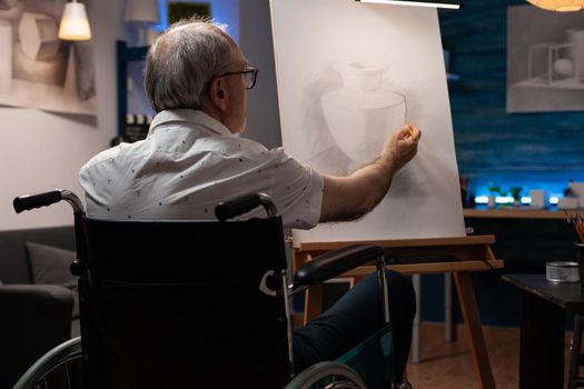 Senior artist with disability drawing vase design with pencil in artwork room at home. Caucasian man sitting in wheelchair creating masterpiece on white canvas and easel