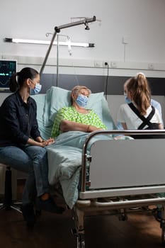 Family with protection face mask against coronavirus sitting with sick grandmother visiting during medical therapy in hospital ward. Retired senior woman resting in bed recovering after surgery