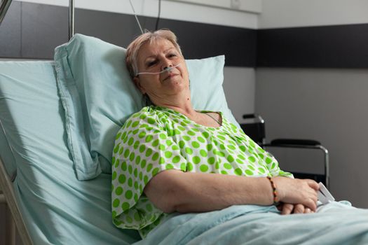 Portrait of sick pensioner woman looking into camera while resting in bed recovering after medical surgery in hospital ward. Hospitalized patient waiting for healthcare treatment during examination
