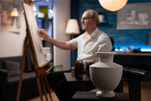 Close up of design of white vase on table with art instruments in artwork studio while elder artist drawing on canvas in background. Aged man working on authentic masterpiece at home