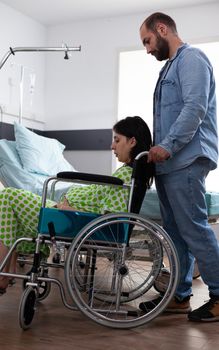 Caucasian man helping woman with pregnancy in wheelchair at medical clinic. Husband supporting pregnant wife while sitting in hospital ward with professional healthcare equipment