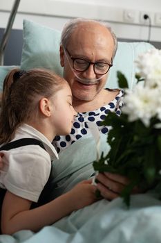 Little granddaughter hugging elderly grandfather visiting him in hospital ward while recovering after medical surgery. Sick senior man patient resting in bed during clinical therapy, heathcare support