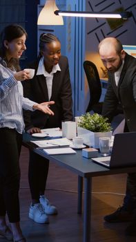 Excited multi ethnic businesspeople receiving good news clapping while standing at table conference overworking in office meeting room late at night. Diverse teamwork looking at company presentation