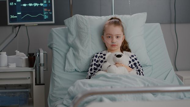 Portrait of lonely little child looking into camera holding teedy bear in hands resting in bed suffering infection. Kid waiting for recovery treatment during medical examination in hospital ward