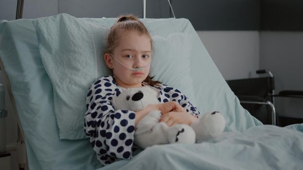 Portrait of little girl patient wearing oxygen nasal tube resting in bed recovery after suffering pneumonia infection. Sad girl having fever during medical consultation in hospital ward