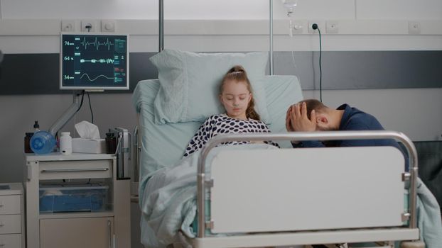 Hospitalized sick daughter sleeping after suffering medical surgery while worried father praying for health recovery. Child with nasal tube resting in bed during sickness examination in hospital ward