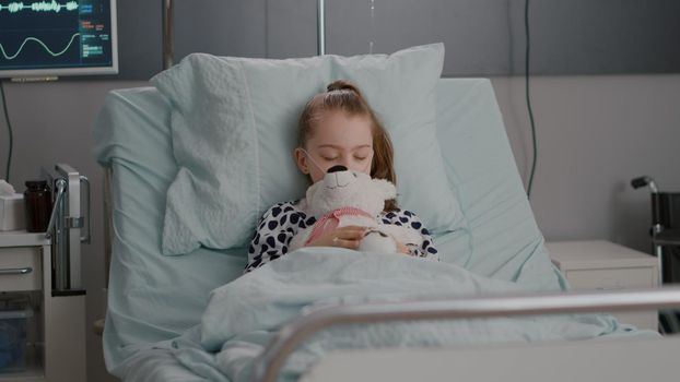 Portrait of tired sick child sleeping after suffering medical recovery surgery during disease examination in hospital ward. Hospitalized kid resting in bed wearing oxygen nasal tube