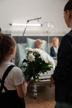 Caring granddaughter bringing flowers gift to sick old grandfather during clinical recovery in hospital ward. Elderly senior man resting in bed recovering after medical surgery. Healthcare support