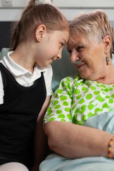 Supporting granddaughter touching forehead with elderly grandmother showing love during clinical recovery, waiting together for healthcare treatment. Sick senior woman resting in bed in hospital ward