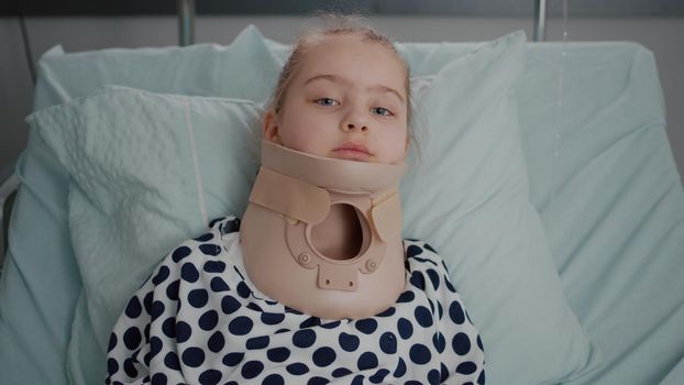 Portrait of sick little patient resting in bed looking at camera while having neck cervical collar recovering after painful surgery in hospital ward. Child wearing oxygen nasal tube during examination