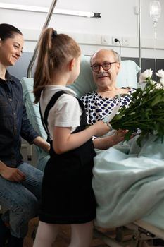 Cheerful granddaughter bringing flowers to elderly grandparent in hospital ward during clinical recovery. Caring family visiting him supporting after medical surgery. Healthcare support service