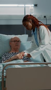 Doctor installing oximeter on patient hand in hospital ward at medical clinic. African american medic doing consultation on sick old man with nasal oxygen tube and IV drip bag for disease