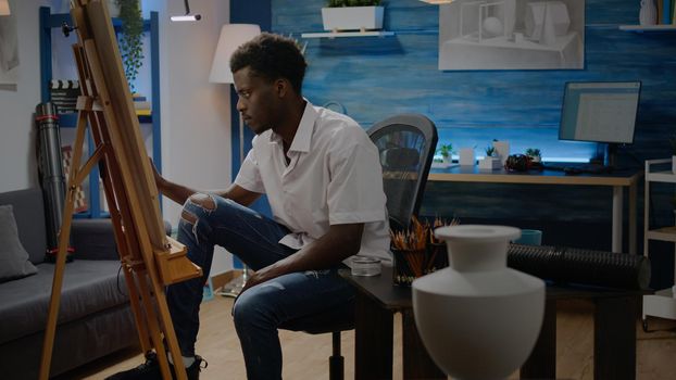 African american adult making fine art sitting in artwork studio with artistic tools. Black young man designing drawing of vase with pencil and white canvas on easel. Person with creativity