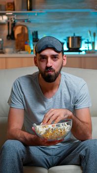 Portrait of man with beard holding popcorn bowl while watching entertainment movie on television. Concentrated person with eye sleep mask sitting on sofa late at night in kitchen