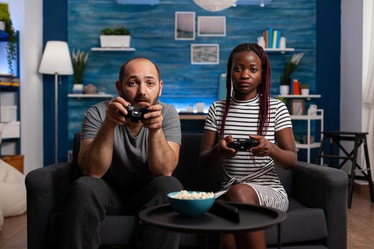 POV of interracial couple playing video game on console holding controllers at television. Multi ethnic partners looking at camera while enjoying fun activity with TV and joysticks